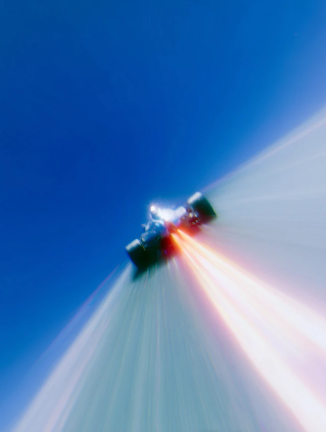 a graphic illustration of a vehicle from wipeout upwards at an angle, characterized by strong lines and color transitions. the vehicle is dark, almost black, with bright orange engine exhaust trails. the backdrop is a clear, smooth gradient of blue at the top, fading to white, and then to a White at the bottom, creating a sense of speed and ascent.